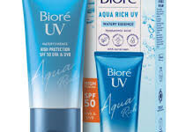 A short flat conical light blue plastic tube with a dark blue plastic lid that has Bioré UV Aqua Water Essence SPF 50 written in bright white writing on it next to a tall rectangular bright white cardboard box that has Bioré UV Aqua Water Essence SPF 50 written in bright white wariting and some pictures of some dark blue stripes on it, on a white background.