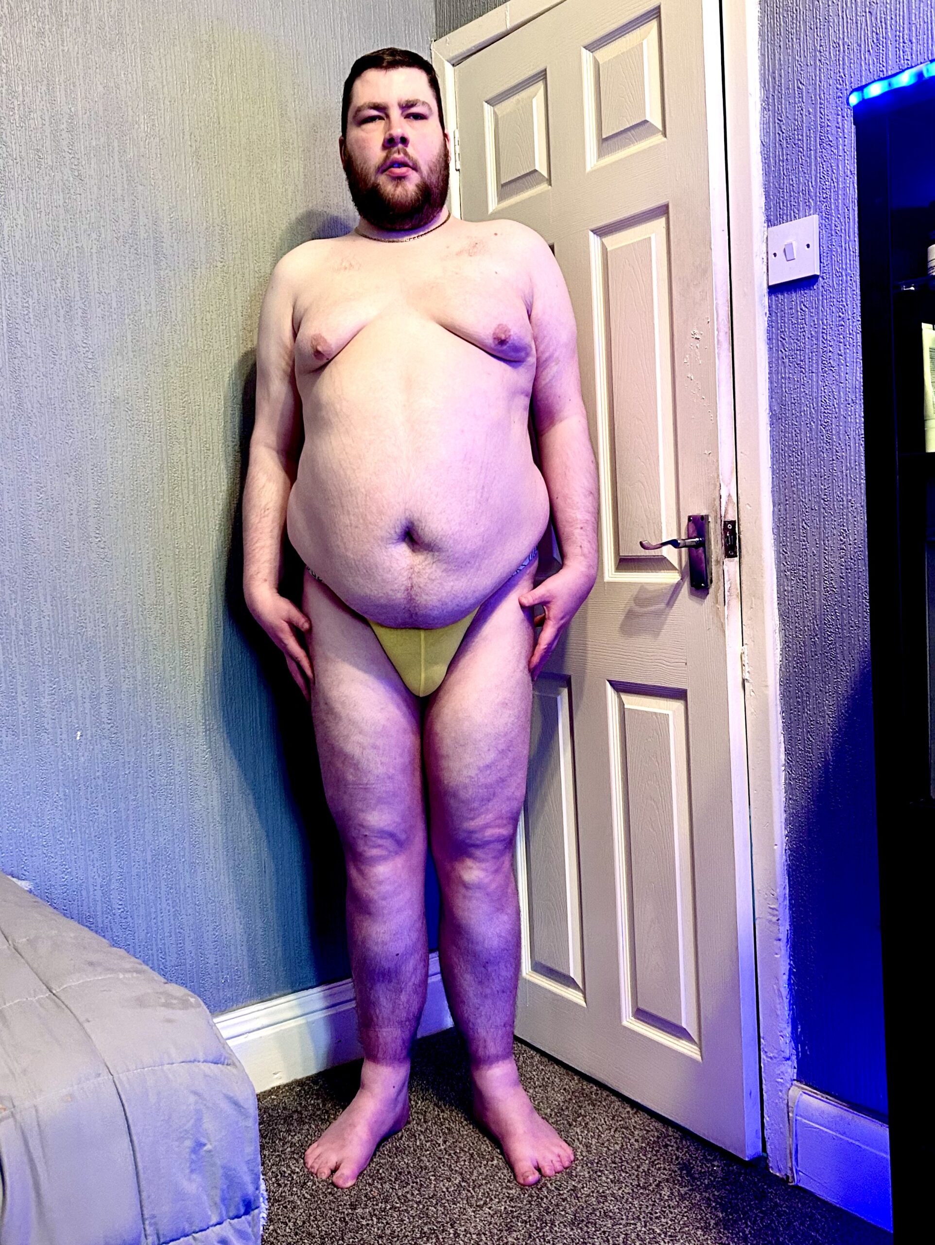 A tall bearded man wearing a bright yellow tanga with a black waistband that has Kiniki written in bright white writing on it stood against a white wooden Door.