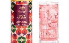 A tall cylindrical bright green, orange and red cardboard tube with a bright gold plastic lid and a bright purple label that has Whittard of Chelsea Sour Cherry Instant Tea written in bright white writing on it next to a tall cylindrical clear glass full of some translucent pink liquid and some cubes of clear ice, on a white background.