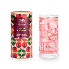 Tea Tastes Terrific | Whittard Of Chelsea Sour Cherry Instant Tea A tall cylindrical bright green, orange and red cardboard tube with a bright gold plastic lid and a bright purple label that has Whittard of Chelsea Sour Cherry Instant Tea written in bright white writing on it next to a tall cylindrical clear glass full of some translucent pink liquid and some cubes of clear ice, on a white background.