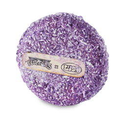 A medium circular dark purple shampoo bar that has some specks of bright white running through it and a white rice paper label that has Lush x Teenage Mutant Ninja Turtles written in bright green writing on it, on a white background.