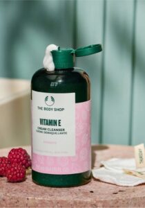 A short cylindrical dark green plastic bottle with a dark green plastic lid and a bright white label that has The Body Shop Vitamin E Cream Cleanser written in dark green writing and some pictures of some dark pink raspberries on it, on a white background.