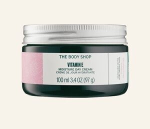 A short cylindrical dark green glass jar with a silver metal lid and a white label that has The Body Shop Moisture Day Cream written in bold black writing, Vitamin E written in dark green writing, and some pictures of some dark pink flowers on it, on a white background.