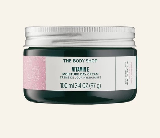 Vitamins Always Help You Get Soothed & Hydrated Skin | The Body Shop Vitamin E Range A short cylindrical dark green glass jar with a silver metal lid and a white label that has The Body Shop Moisture Day Cream written in bold black writing, Vitamin E written in dark green writing, and some pictures of some dark pink flowers on it, on a white background.