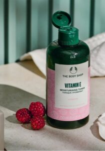 A tall cylindrical dark green plastic bottle with a dark green plastic lid and a bright white label that has The Body Shop Vitamin E Moisturising Toner written in dark green writing and some pictures of some dark pink raspberries on it, on a white background.