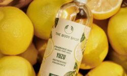 A tall cylindrical clear glass bottle full of some clear liquid with a silver metal lid and a bright yellow label that has The Body Shop Yuzu Fragrance Mist written in dark green writing, some pictures of some large bright yellow yuzus, and some pictures of some dark green leaves on it, on a white background.