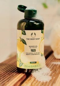 A tall cylindrical dark green plastic Bottle with a dark green plastic lid and a bright yellow label that has The Body Shop Yuzu Shower Gel written in bold black writing, some pictures of some large bright yellow Yuzus, and some pictures of some dark green leaves on it, on a white background.