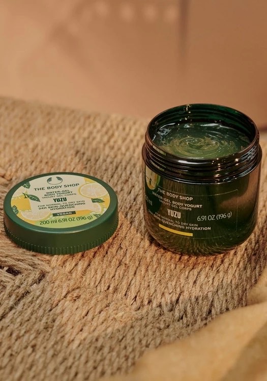 A short rounded rectangular dark green plastic tub with a dark green plastic lid and a bright Yellow label that has The Body Shop Yuzu Water-Gel Body Yoghurt written in bold black writing, some pictures of some large bright yellow Yuzus, and some pictures of some dark green leaves on it, on a white background.