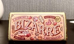 A large rectangular chocolate bar wrapped in a light pink paper wrapper that has The Chocolate Smiths written in bold black writing, Ice Cream Twist written in dark red writing, Bizarre Bar written in bright white writing and some pictures of a Pineapple, a Strawberry, a Beach Ball, an Ice Cream Cone, an Ice Lolly, a Flamingo Pool Float and a Slice of Lime on it, on a dark wooden cabinet.