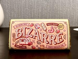A large rectangular chocolate bar wrapped in a light pink paper wrapper that has The Chocolate Smiths written in bold black writing, Ice Cream Twist written in dark red writing, Bizarre Bar written in bright white writing and some pictures of a Pineapple, a Strawberry, a Beach Ball, an Ice Cream Cone, an Ice Lolly, a Flamingo Pool Float and a Slice of Lime on it, on a dark wooden cabinet.