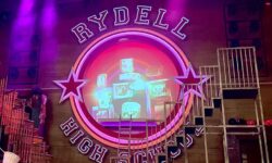 A large rectangular dark wooden stage that has a large circular light wooden sign that has Rydell High School written in bright white writing and some pictures of some old fashioned TVs on it in the middle of it, with a bright red Star either side of it that's surrounded by some bright silver metal staircases and some bright pink and purple lights.