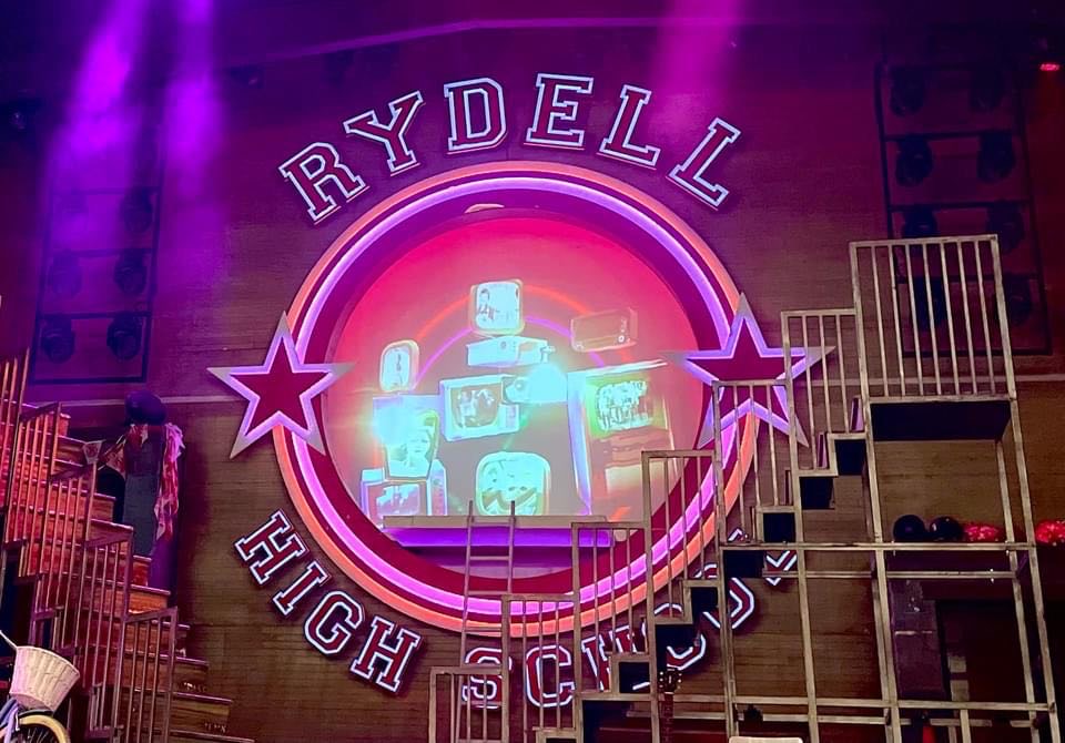 A large rectangular dark wooden stage that has a large circular light wooden sign that has Rydell High School written in bright white writing and some pictures of some old fashioned TVs on it in the middle of it, with a bright red Star either side of it that's surrounded by some bright silver metal staircases and some bright pink and purple lights.
