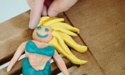 A hand holding a large Mermaid shaped block of Plasticine that has a bright pink face, some bright yellow hair, and a bright turquoise tail, on a white background.