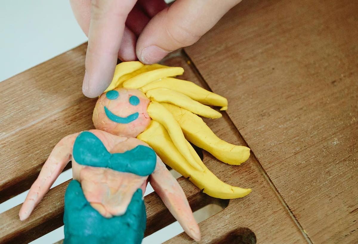 A hand holding a large Mermaid shaped block of Plasticine that has a bright pink face, some bright yellow hair, and a bright turquoise tail, on a white background.