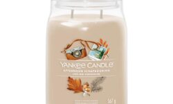 A tall cylindrical glass jar full of some pale brown wax with a label that has Yankee Candle written in bright white writing, Afternoon Scrapbooking written in bright white writing, and a picture of a small vintage camera, a postcard, some dark brown autumn leaves, and some dark brown acorns on it, on a white background.