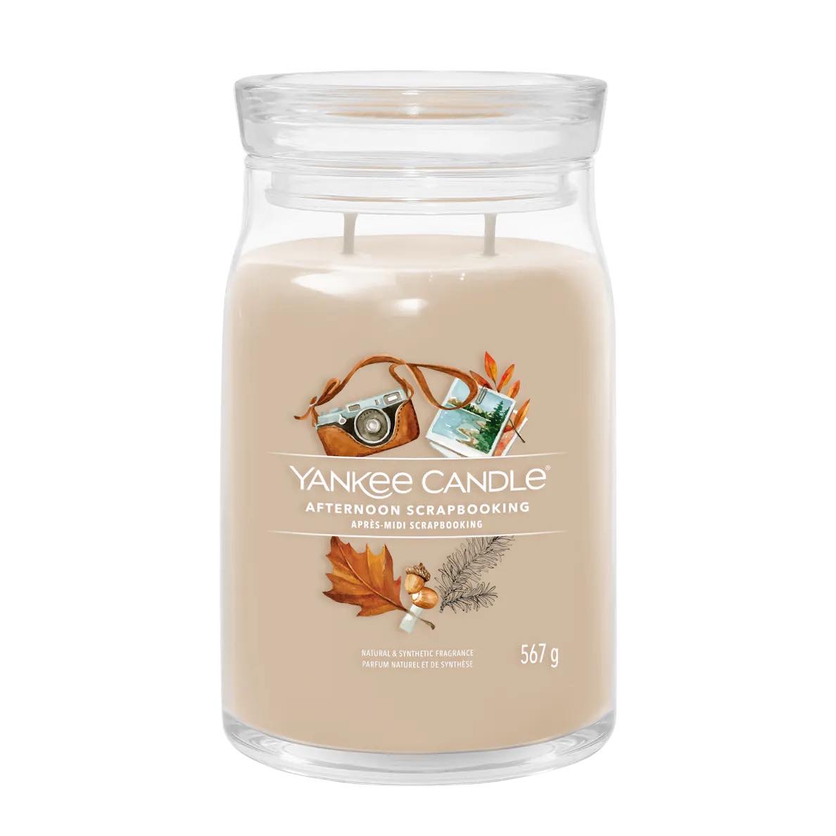 I'm Getting Away To A Cosy Cottage This Autumn | Yankee Candle Cottage Getaway Collection A tall cylindrical glass jar full of some pale brown wax with a label that has Yankee Candle written in bright white writing, Afternoon Scrapbooking written in bright white writing, and a picture of a small vintage camera, a postcard, some dark brown autumn leaves, and some dark brown acorns on it, on a white background.