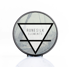 A short cylindrical silver metal tin with a silver metal lid and a gradient dark grey label that has Runesilk Elements Earth Beard Butter written in bold black writing and a picture of a bright white triangle on it, on a white background.
