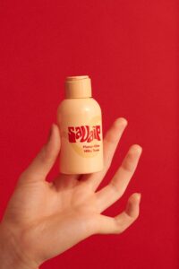A short cylindrical light peach plastic bottle with a light peach plastic pump lid and a gradient bright orange and red label that has Savaip Plump Glow Milky Toner written in bright red writing on it, on a white background.
