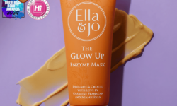 A tall conical bright orange plastic tune with a bright orange plastic lid that has Ella and Jo The Glow Up Enzyme Mask written in bright white writing and some pictures of some pale orange flowers and leaves on it, on a pale lilac background.