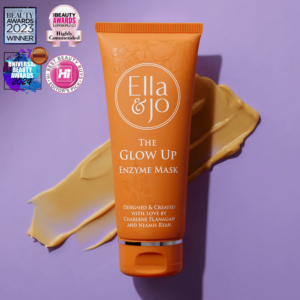A tall conical bright orange plastic tune with a bright orange plastic lid that has Ella and Jo The Glow Up Enzyme Mask written in bright white writing and some pictures of some pale orange flowers and leaves on it, on a pale lilac background.