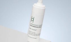 A tall cylindrical bright white plastic bottle with a bright white plastic lid and a bright white label that has Harrogate Organics Magnesium Spray written in bright green writing and a picture of two overlapping bright green letter h’s on it, on a white background.