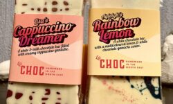 A small rectangular bright white chocolate bar that has some light brown spots all over it with a gradient orange and pink label that has La Chocolatrice written in bright pink writing and The Cappuccino Dreamer Chocolate Bar written in bold black writing on it next to a small rectangular bright white chocolate bar that has some bright blue and pink speckles all over it with a gradient bright orange and yellow label that has La Chocolatrice written in bright pink writing and The Rainbow Lemon Chocolate Bar written in bold black writing on it, on a white background.