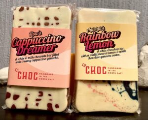 A small rectangular bright white chocolate bar that has some light brown spots all over it with a gradient orange and pink label that has La Chocolatrice written in bright pink writing and The Cappuccino Dreamer Chocolate Bar written in bold black writing on it next to a small rectangular bright white chocolate bar that has some bright blue and pink speckles all over it with a gradient bright orange and yellow label that has La Chocolatrice written in bright pink writing and The Rainbow Lemon Chocolate Bar written in bold black writing on it, on a white background.
