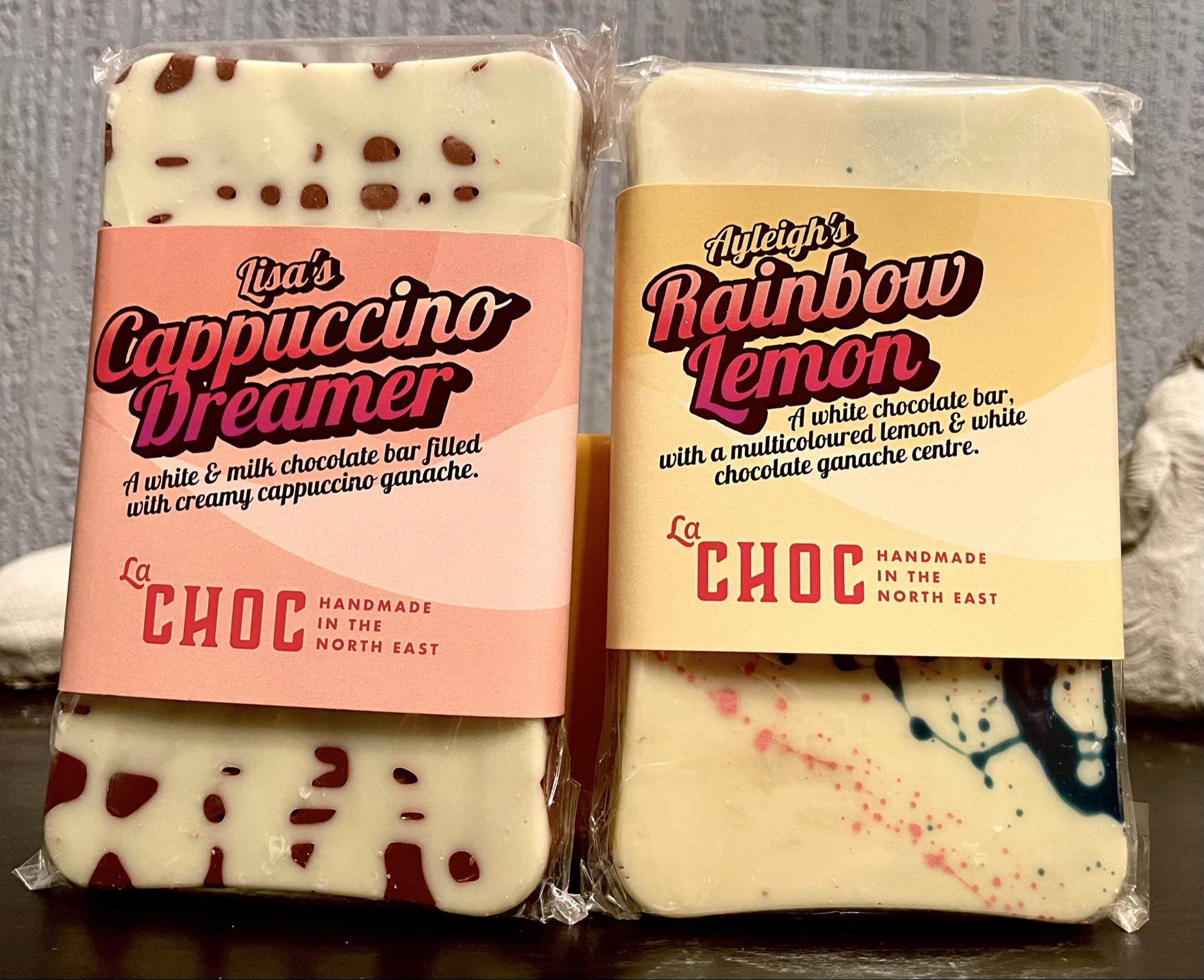 A small rectangular bright white chocolate bar that has some light brown spots all over it with a gradient orange and pink label that has La Chocolatrice written in bright pink writing and The Cappuccino Dreamer Chocolate Bar written in bold black writing on it next to a small rectangular bright white chocolate bar that has some bright blue and pink speckles all over it with a gradient bright orange and yellow label that has La Chocolatrice written in bright pink writing and The Rainbow Lemon Chocolate Bar written in bold black writing on it, on a white background.