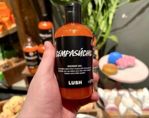 A hand holding a tall cylindrical clear plastic bottle full of some shimmering bright orange shower gel with a black label that has Lush Cempasúchil Shower Gel written in bold white writing on it in front of a large rectangular light wooden table.