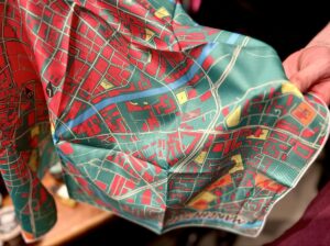 A hand holding a large rectangular piece of crumpled up bright green fabric that has a bright red, yellow, green and blue map of Manchester pattern all over it.