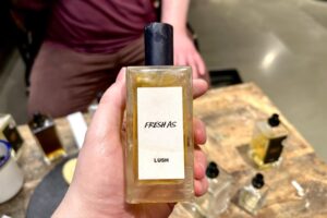 A hand holding a tall rectangular clear glass perfume bottle full of some amber coloured liquid with a metallic black lid and a white label that has Lush Fresh As Perfume written in bold black writing on it in front of a large rectangular light wooden table.