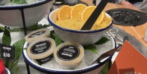 A large circular white plastic bowl with a dark blue rim full of some golden yellow face mask that has some slices of orange on top of it and a black wooden spatula sticking out of the top of it next to several short cylindrical clear plastic tubs with black labels that all have Lush Turmeric Fresh Face Mask written in bold bright white writing on them on top of a pile of some clear crushed ice on a light wooden table.