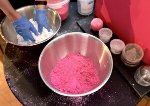 A large circular black metal Bowl full of some bright white powder that has a pair of hands wearing a pair of bright blue rubber gloves over it next to a large circular black metal bowl full of some bright pink powder, a pile of some clear plastic tubs that all have Lush written in bold black writing on them, and a medium gradient bright pink and white rounded hexagonal shaped bath bomb on a light wooden table.