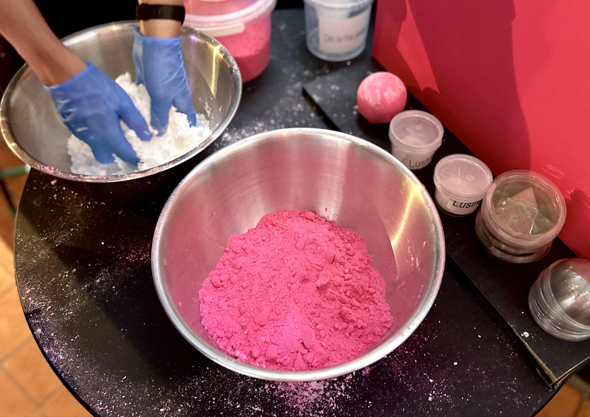 Marvellous Marzipan Magic | Lush Snowcake Soap and Snow Angel Bath Melt A large circular black metal Bowl full of some bright white powder that has a pair of hands wearing a pair of bright blue rubber gloves over it next to a large circular black metal bowl full of some bright pink powder, a pile of some clear plastic tubs that all have Lush written in bold black writing on them, and a medium gradient bright pink and white rounded hexagonal shaped bath bomb on a light wooden table.