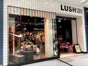 A large rectangular brightly lit glass store front with a large rectangular black board over its door that has Lush written in bold white writing on it, on a white background.