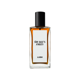 A short rectangular glass perfume bottle full of some translucent amber liquid with a metallic black lid and a white label that has Lush The Bee’s Knees Perfume written in bold black writing on it, on a white background.