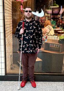 A tall bearded man wearing a pair of rectangular silver metal sunglasses with some dark brown lenses, a black long sleeve shirt that has a white and red moose print all over it, a dark red V Neck t-shirt, a pair of dark red chinos and a pair of shiny red shoes holding a bright white cane with a red leather handle stood in front of a large rectangular shop window.