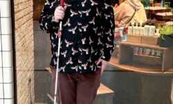 A tall bearded man wearing a pair of rectangular silver metal sunglasses with some dark brown lenses, a black long sleeve shirt that has a white and red moose print all over it, a dark red V Neck t-shirt, a pair of dark red chinos and a pair of shiny red shoes holding a bright white cane with a red leather handle stood in front of a large rectangular shop window.