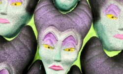 A pile of some large glittery bright green woman’s head shaped bath bombs that has some bright purple eyeshadow, some bright yellow eyes, some bright pink lips, and some dark purple horns on them, on a bright lime green background.