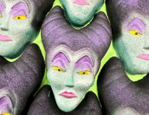 A pile of some large glittery bright green woman’s head shaped bath bombs that has some bright purple eyeshadow, some bright yellow eyes, some bright pink lips, and some dark purple horns on them, on a bright lime green background.