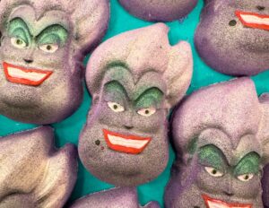A pile of some large glittery bright purple Sea Witch’s Head shaped bath bombs that has some bright green eyeshadow, some bright red lipstick, some dark purple eyebrows, and a wide mischievous grin engraved on them, on a bright turquoise background.