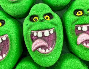 A pile of some large bright green ghost’s head shaped bath bombs that has some bright yellow eyes, some bright white teeth, and and a bright pink tongue on them, on a dark green background.