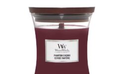 A tall hourglass glass jar full of some dark red wax with a dark wood lid and a bright white label that has WoodWick Candles Phantom Cherry written in bold black writing on it, on a white background.