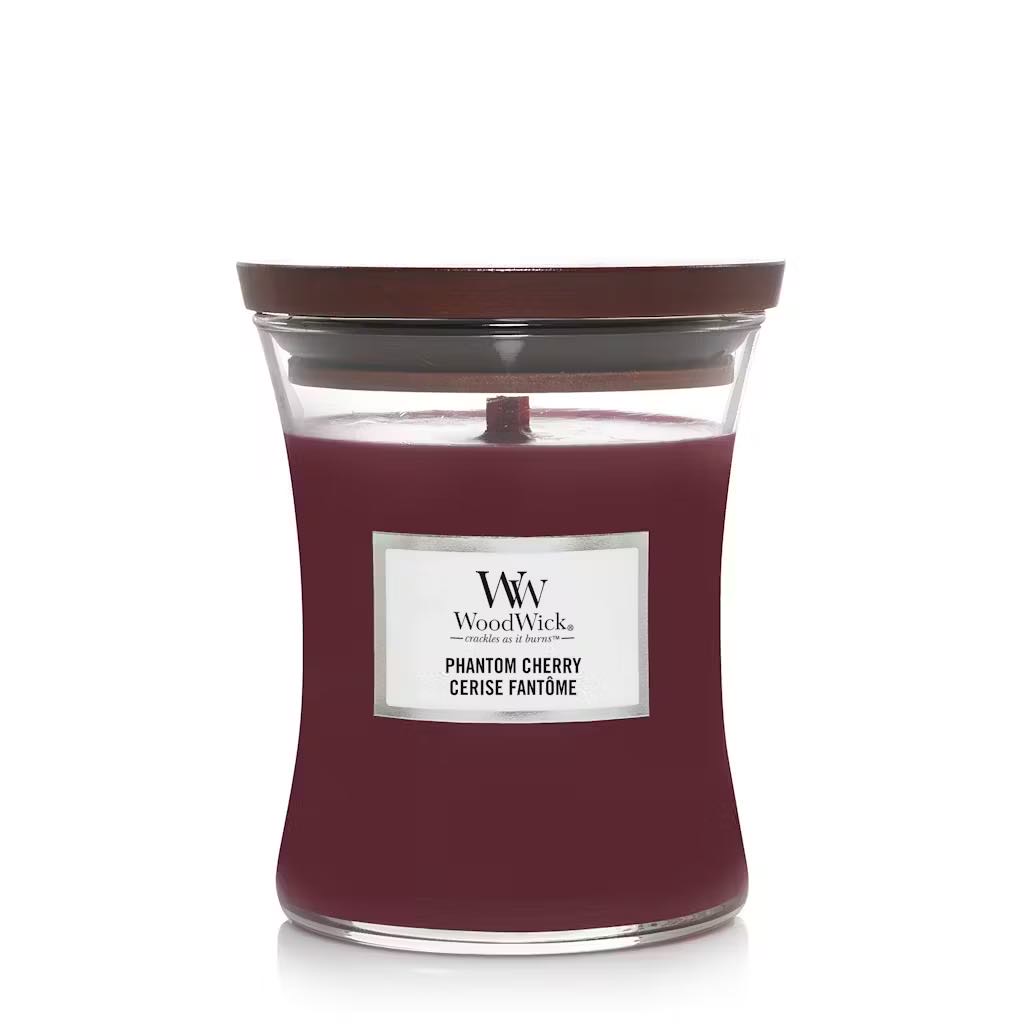 A tall hourglass glass jar full of some dark red wax with a dark wood lid and a bright white label that has WoodWick Candles Phantom Cherry written in bold black writing on it, on a white background.