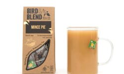 A large rectangular brown cardboard box that has Bird & Blend Tea Company Mince Pie Teabag Gift written in bold black writing and a cut out of a large rectangle on it next to a short cylindrical clear glass mug full of some dark brown liquid, on a white background.