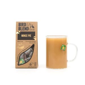 A large rectangular brown cardboard box that has Bird & Blend Tea Company Mince Pie Teabag Gift written in bold black writing and a cut out of a large rectangle on it next to a short cylindrical clear glass mug full of some dark brown liquid, on a white background.