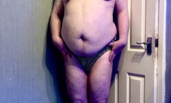 A tall bearded man with his Hands on his Hips wearing a Black Brief with a bright white wavy concentric circle pattern all over it and a black Waistband that has DWNLOW Menswear written in bright white writing on it stood against a white wooden Door.