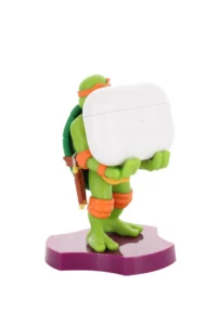 A tall bright green Teenage Mutant Ninja turtle figurine that has a bright orange mask, a bright orange belt, some bright orange elbow pads, and some bright orange knee pads holding a small rectangular bright white earphones case stood on a bright purple turtle shell, on a white background.