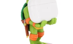 A tall bright green Teenage Mutant Ninja turtle figurine that has a bright orange mask, a bright orange belt, some bright orange elbow pads, and some bright orange knee pads holding a small rectangular bright white earphones case stood on a bright purple turtle shell, on a white background.
