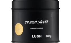 A short cylindrical metallic black Tin with a metallic black lid and a black label that has Lush 29 High Street Single Wick Candle written in bright white writing and Community written in bright white writing inside a bright gold circle on it, on a white background.